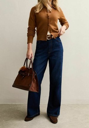 Vrouw draagt een bruin vest met knopen, wijd uitlopende donkerblauwe spijkerbroek, bruine mocassinschoenen, en houdt een bruine suède handtas met pompon-accessoires vast.