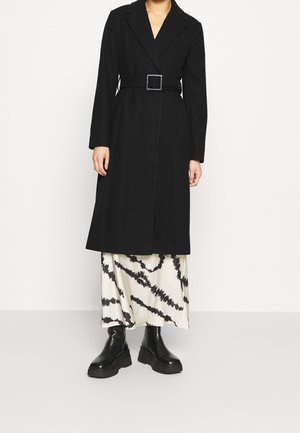 Person trägt einen langen schwarzen Mantel mit Gürtel über einem weißen und schwarzen Batik-Rock, kombiniert mit schwarzen klobigen Stiefeletten.