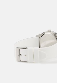 Hvit silikonerem med sølv metallspenne, som har en glatt tekstur og et minimalistisk design. En del av det runde klokkehuset er synlig.