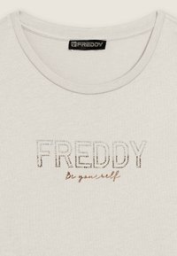 Maglietta grigia chiaro con colletto rotondo, caratterizzata dalla scritta "FREDDY" in strass e la frase "Essere te stesso" in un carattere corsivo sotto. Texture morbida.