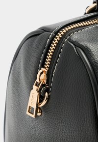 Schwarze, strukturierte Lederhandtasche mit einem Reißverschluss in Goldtone, auffälligen Nähdetails und einem markierten Zipper-Puller. Nahaufnahme des Reißverschlussbereichs.