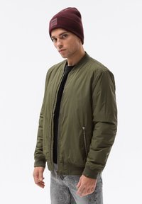 Giacca bomber verde oliva realizzata in materiale morbido con zip frontale, tasche laterali e polsini a coste. Indossata con un berretto di lana bordeaux.