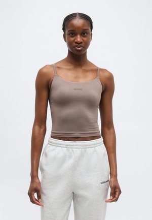 Młoda kobieta z zaplecionymi włosami, ubrana w dopasowany beżowy top na ramiączkach i jasnoszare spodnie dresowe z elastycznym pasem.