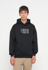Μαύρος φούτερ με κουκούλα από παχύ ύφασμα, διαθέτει μπροστινή τσέπη και λευκό κείμενο "VISION" με επικαλυπτόμενα γράμματα. Χαλαρή εφαρμογή.