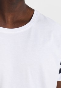 Nærbillede af en person iført en hvid t-shirt med rund hals og detaljer med sorte og hvide striber på ærmerne, sat mod en ensfarvet baggrund.