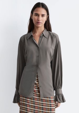 Kobieta w szarej, długorękawowej bluzce z guzikami i falbanianymi mankietami oraz kolorowej, wielokolorowej spódnicy w kratę o wysokim stanie, stojąca na białym tle.