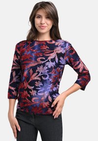 Samt-Pullover mit Dreiviertel ärmeln, der ein florales Muster in Rot und Lila auf einem dunkelblauen Hintergrund zeigt. Weiche Textur.