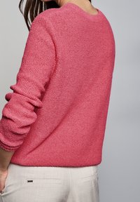 Rosa stickad tröja med en texturerad yta, med långa ärmar och rundad halsringning, bar över ljusa byxor.