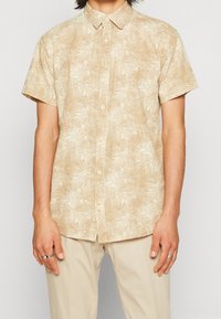 Chemise beige à manches courtes avec un motif de feuilles de palmier, design à boutons, et boutons clairs ; portée avec un pantalon de couleur claire.