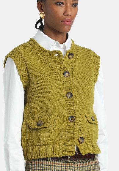 Femme portant un gilet en tricot épais vert olive à boutons sur une chemise blanche et un pantalon à carreaux, accessoirisé avec des boucles d'oreilles en forme de cœur en or.