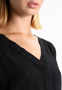 Top negro de cuello en V hecho de una tela suave, con un corte holgado y mangas cortas. El escote presenta detalles de costura limpios y discretos.