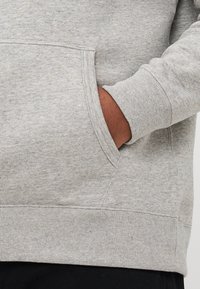 Hånd inne i frontlommen på en lys grå hettegenser med ribbestrikkede mansjetter og kant, brukt med svarte bukser.