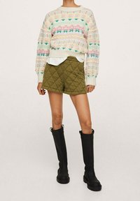 Pull en tricot avec motifs pastel, short matelassé vert olive, et bottes en caoutchouc noires montant jusqu'aux genoux. Le tissu texturé crée un contraste de couleurs.