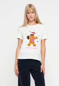 Vrouw in een wit T-shirt met een peperkoekman in zonnebril, kerstmanhoed, roze tas, drankje en de tekst "Hier ben ik eruitzien als een snack."