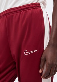Άτομο που φοράει μπορντό αθλητικές φόρμες Nike με λευκές πλαϊνές ρίγες και εμφανές χέρι που ακουμπάει δίπλα στο σώμα.