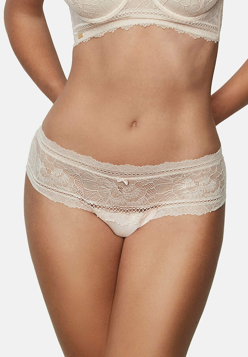 Culotte hipster en dentelle de couleur nude, ornée de motifs floraux, avec des bords festonnés et un petit nœud au centre devant. Matière douce et extensible.