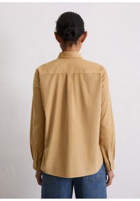 Long-sleeve, tan corduroy shirt with a back yoke, button cuffs, and a straight hem, paired with blue jeans, viewed from the back.