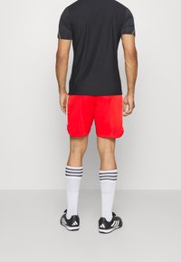 Pantaloni sportivi rossi abbinati a una maglietta nera. I pantaloni presentano una texture liscia e l'outfit include scarpe nere con dettagli bianchi e calze a righe.