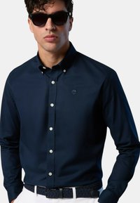 Camicia button-up blu navy con una texture liscia, caratterizzata da un piccolo logo e bottoni bianchi, indossata con una cintura scura a trama.