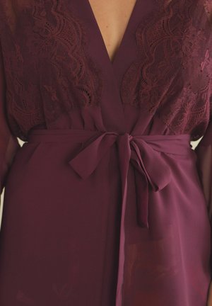 Bordeaux jurk met een kanten overlay in een golvend patroon, een diepe V-hals en een strikdetail in de taille. Doorzichtig materiaal voegt textuur toe.