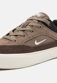 Smeđi nubuk tenisice s bijelim Nike logotipom, teksturiranom tkaninom, ravnim vezicama i bijelim gumiranim potplatom s teksturiranim uzorkom.