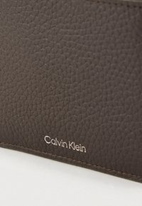 Cartera de cuero marrón texturizado con superficie en relieve y costuras sutiles, con un logo plateado "Calvin Klein" en el frente.