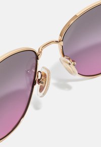 Ochelari de soare tip cat-eye cu ramă din metal roz aurie și lentile violet cu degradé. Prezintă pernuțe nazale transparente și un design modern și elegant.