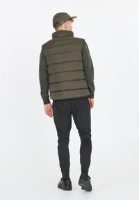 Olivengrüne gepolsterte Weste mit hohem Kragen, kombiniert mit einem dunkelgrauen Langarmshirt und schwarzen Slim-Fit-Hosen, von hinten betrachtet.