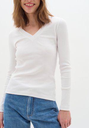 Jeune femme souriante, portant un haut blanc ajusté à manches longues côtelé avec col en V et un jean bleu sur un fond blanc uni.