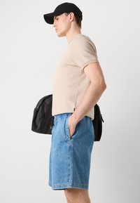 T-shirt beige texturé, shorts en denim bleu, casquette noire, tenant un sac à dos noir, vue de côté d'un modèle en tenue décontractée.