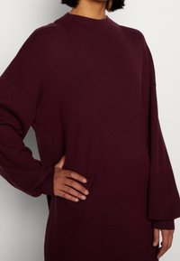 Burgunderfarbener oversized Pullover mit hohem Kragen, weit geschnittenen Ärmeln und gerippten Bündchen. Glatte Textur, minimalistisches Design und keine sichtbaren Muster oder Akzente.