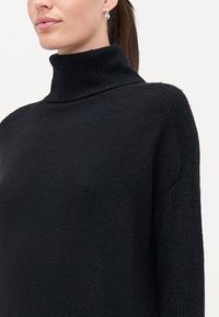 Melnas turtlenecka tipa džemperis ar strukturētu adījumu, augstu apkakli un krituma plecu dizainu, izgatavots no mīksta, bieza materiāla.