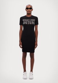 Čierne priliehavé šaty s krátkymi rukávmi, na hrudníku s textom "VERSACE JEANS COUTURE" v striebornej farbe. Skombinované s bielymi teniskami.