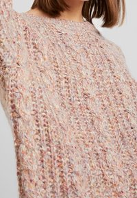 Gros plan sur une personne portant un pull en tricot épais multicolore rose et beige avec un col rond et un motif texturé.