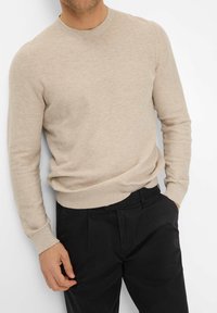 Maglione a maglia beige con girocollo, maniche lunghe e polsini a coste, abbinato a pantaloni neri, che mostrano una vestibilità comoda e una texture interessante.