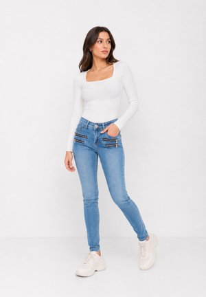 Femme portant un haut blanc à manches longues avec un décolleté carré, un jean bleu clair avec plusieurs fermetures éclair à l'avant, et des baskets blanches épaisses, debout devant un fond blanc.