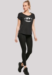 Svart t-skjorte med korte ermer og stort Batman-logo, kombinert med svarte leggings og svarte treningsko; glatt stoff med en avslappet passform.