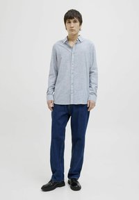 Camisa de botones a rayas azul claro y blanca con cuello, combinada con unos vaqueros rectos azul oscuro y zapatos negros, de pie frente a un fondo neutro.