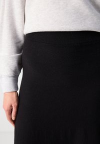 Zwarte gebreide rok met een geribde tailleband, gecombineerd met een lichtgrijze sweatshirtmouw, die een soepele textuur en een aansluitend ontwerp laat zien.