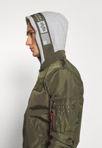 Olivgrüner Bomberjacke mit grauem Kapuzen. Verfügt über einen Reißverschluss, gerippte Bündchen und mehrere Taschen. Branding auf dem Kapuzeneinsatz.