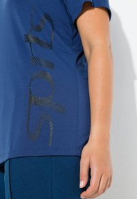 Tmavě modré tričko z hladké látky s texturovaným černým hadovitým vzorem na boku. Volný střih, krátké rukávy.