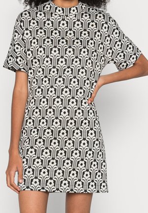 Frau trägt ein schwarz-weiß geblümtes Kleid mit kurzen Ärmeln, eine Hand in der Hüfte, neutraler Hintergrund.