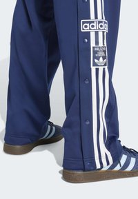 Pantalones deportivos marinos con franjas laterales blancas, botones a presión y un parche con logo; combinados con zapatillas a rayas en azul marino y celeste.