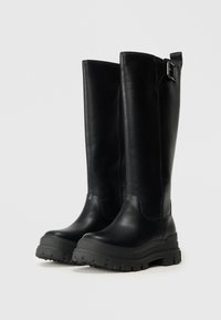 Fekete bőr, térd fölé érő csizmák matt felülettel, lapos gumitalppal és oldalra rögzített csattal. Masszív, textúrált talppal rendelkezik.