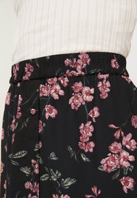 Blommig svart tygkjol med elastisk midja, prydd med rosa blommor och gröna löv, strukturerad i en mjuk, flytande design.