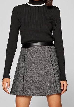 Femme portant un haut côtelé noir à manches longues avec un bord blanc au col, rentré dans une jupe chevron gris taille haute avec une ceinture noire.