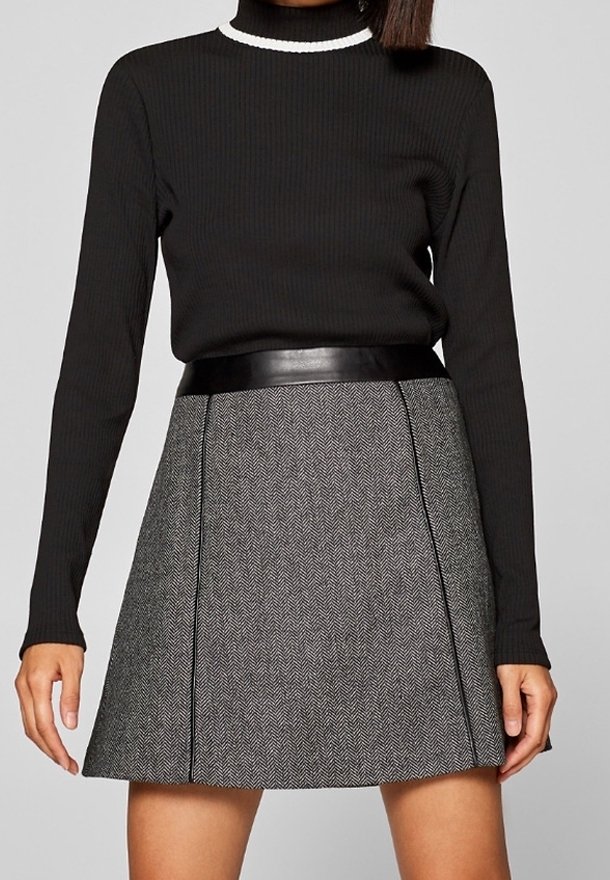 Femme portant un haut côtelé noir à manches longues avec un bord blanc au col, rentré dans une jupe chevron gris taille haute avec une ceinture noire.