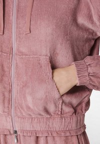 Chaqueta de pana rosa con cremallera y capucha, que presenta una textura acanalada, puños elásticos y un bolsillo frontal.