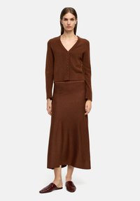 Brązowy sweter z dzianiny z dekoltem w serek i zapięciem na guziki, w połączeniu z pasującą długą brązową spódnicą. Modelka nosi brązowe buty wsuwane.