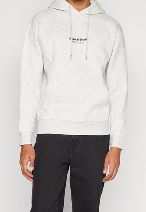 Osoba w jasno szarej bluzie z kapturem z czarnym napisem na środku oraz czarnych spodniach, stojąca na jednolitym tle.